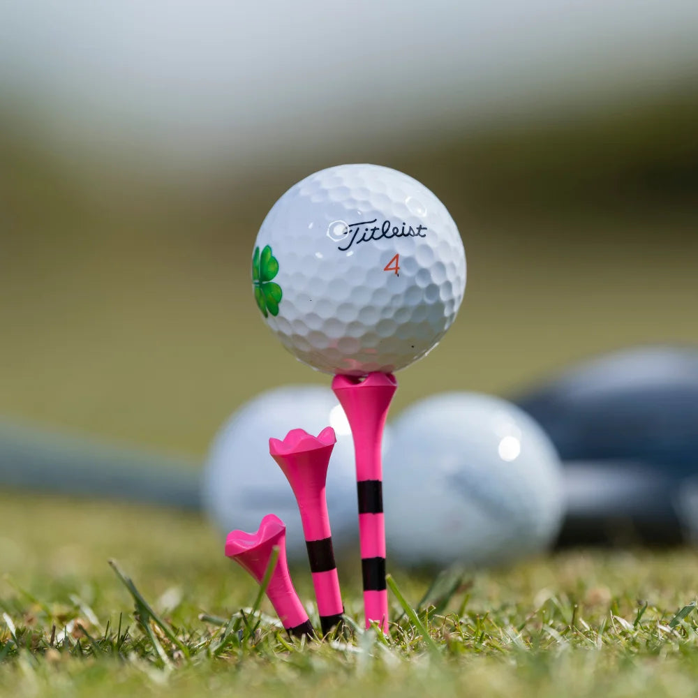 A titleist golf ball with a clover imprinted on the ball while positioned on top of a citrus pink striped pride performance golf tee.