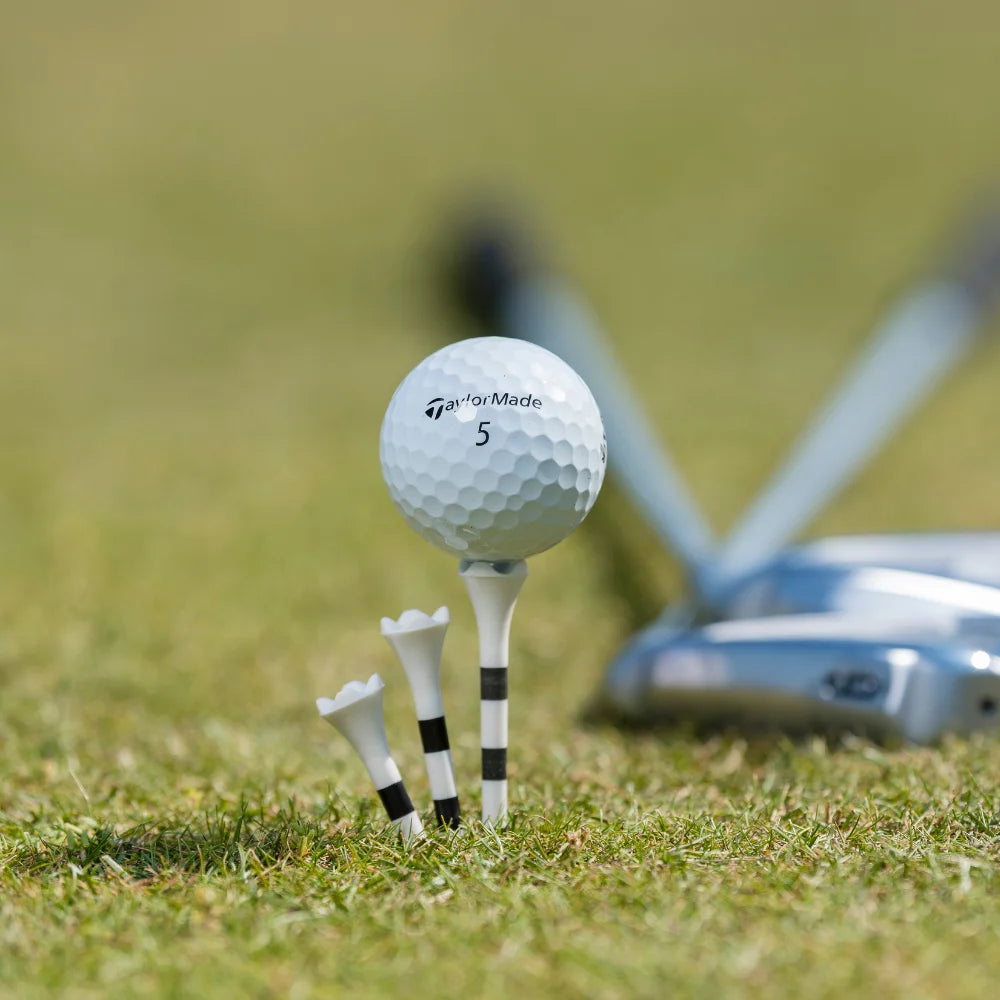 A TaylorMade golf ball positioned on top of a white and black striped pride performance tee with golf clubs behind it.