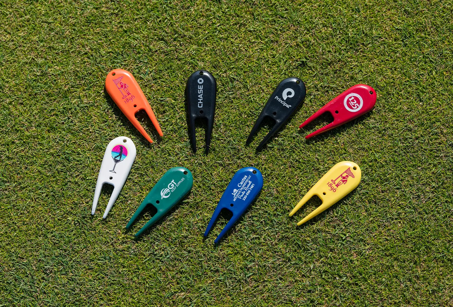 Eight multi-colored custom divot repair tools with different imprinted logos on them positioned on green turf.