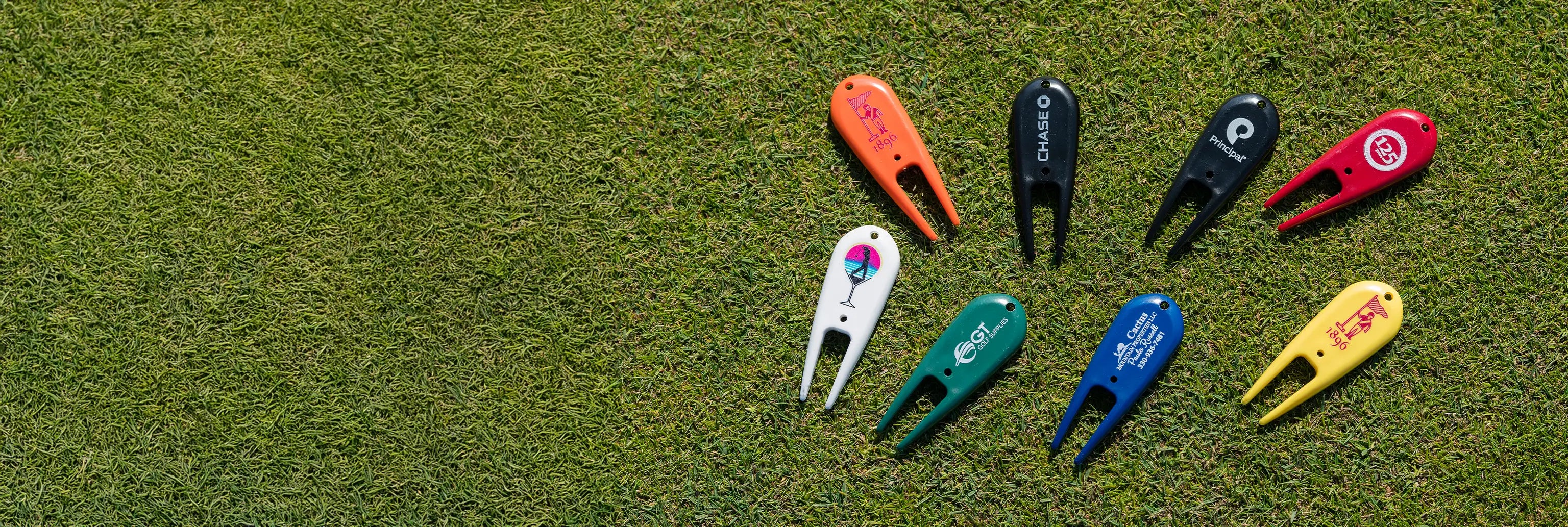 Eight multi-colored custom divot repair tools with different imprinted logos on them positioned on green turf.