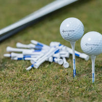 Two taylormade golf balls on tees beside a pile of white and blue golf tees on grass.