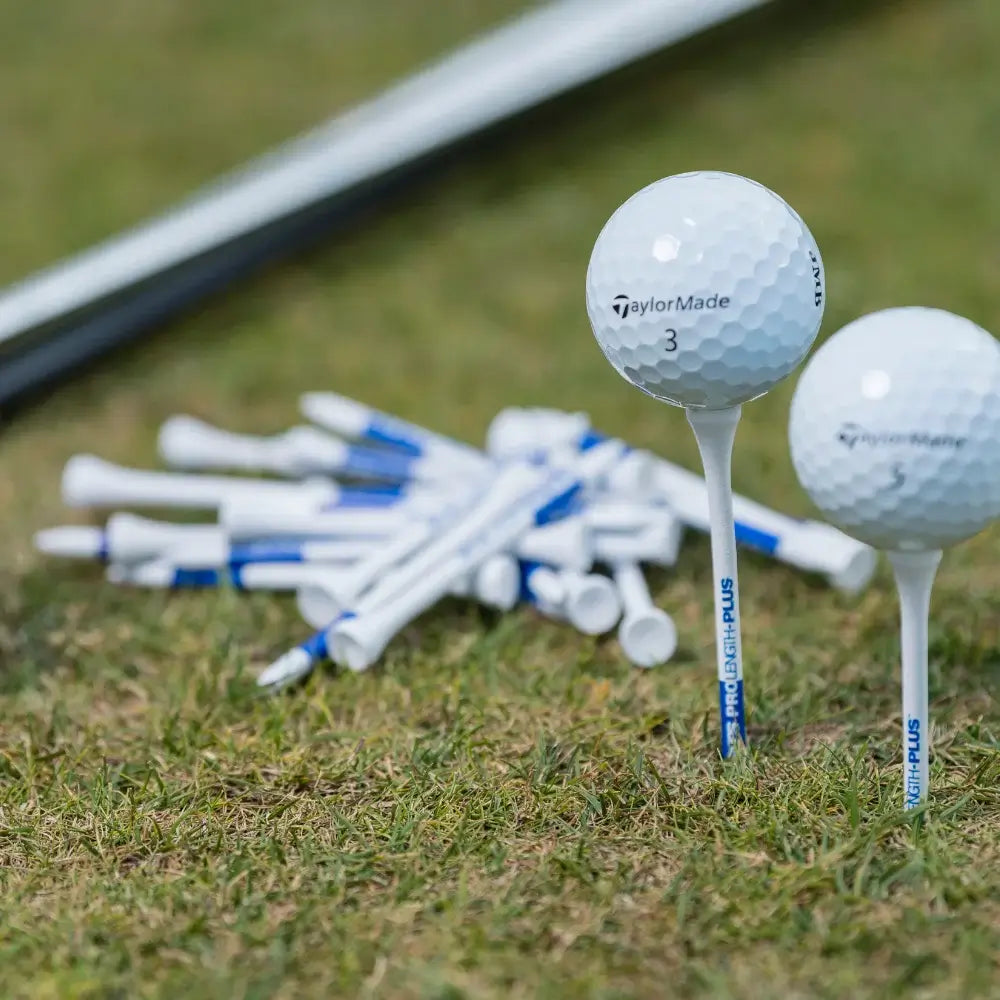 Two taylormade golf balls on tees beside a pile of white and blue golf tees on grass.