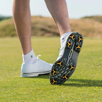 A close-up of a golfer's foot in a white shoe with scorpion stinger golf spikes, standing on green grass.