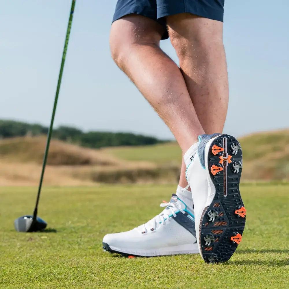 A close-up of a golfer's foot wearing a white golf shoe and silver tornado golf spikes on a grassy golf course, with a golf club visible in the background.