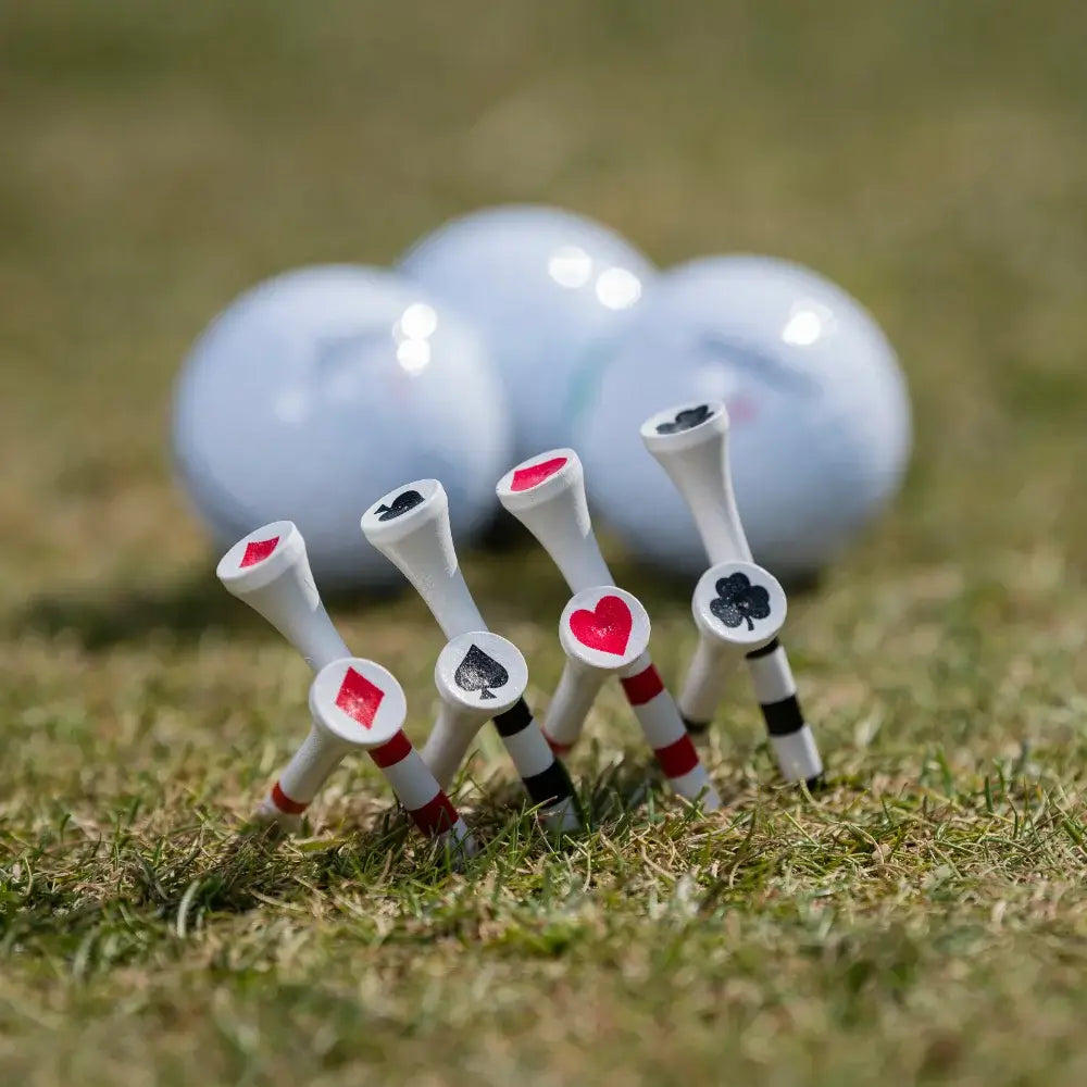 A close-up of golf tees with playing card symbols, and golf balls in the background on a grassy surface.