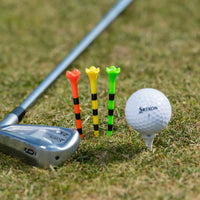A golf club, three colorful tees (red, yellow, and green with black stripes), and a white srixon golf ball on a tee on grass.