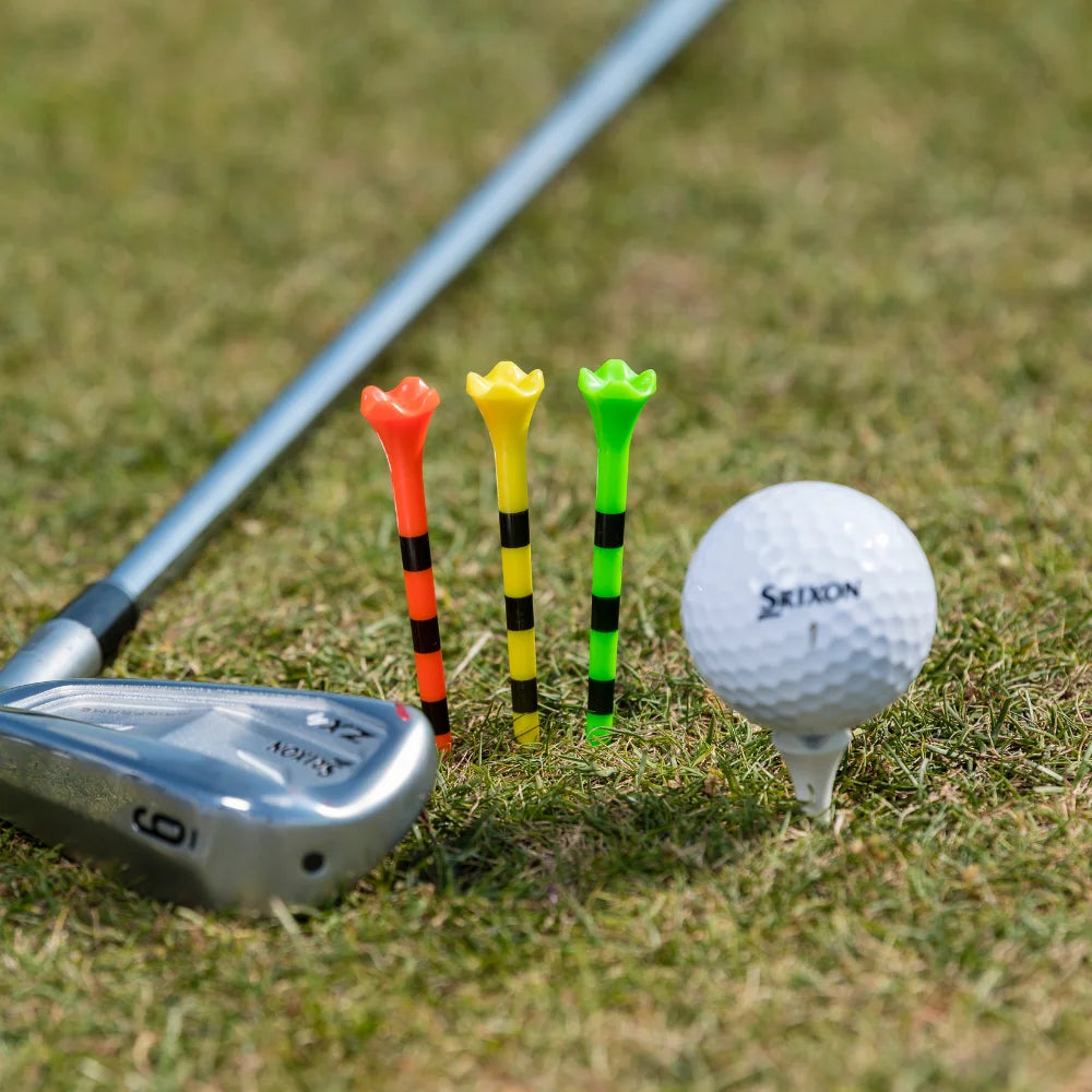 A golf club, three colorful tees (red, yellow, and green with black stripes), and a white srixon golf ball on a tee on grass.