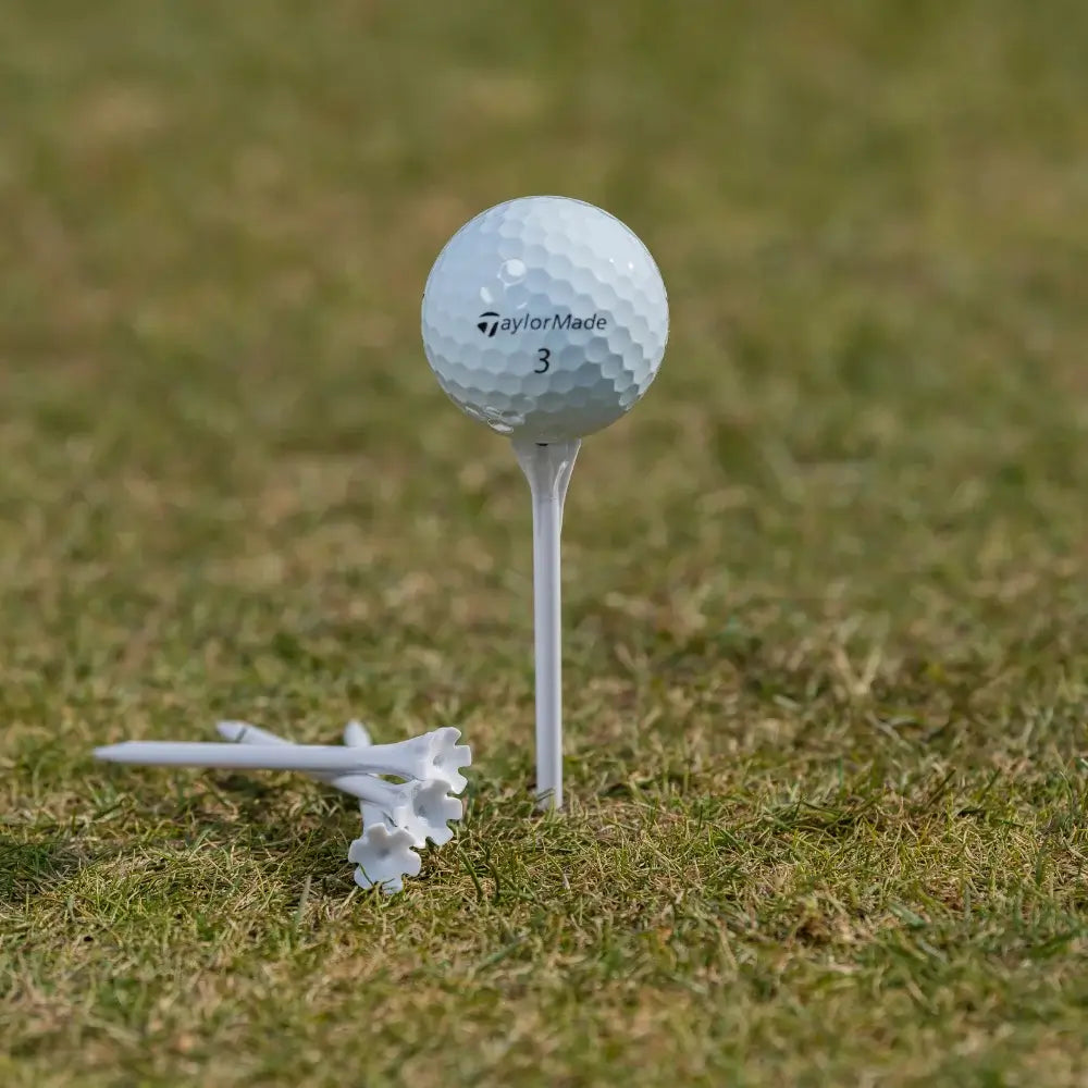 A white taylormade golf ball on a white tee, with additional white tees in the foreground, set on grassy ground.