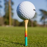 A golf ball rests atop a 3 1/4-inch bamboo golf tee with teal, red, orange, and green sunrise gradient stripes, on grass.