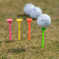 A taylormade golf ball on a green tee, with three brightly colored tees (pink, yellow, and orange) in front.