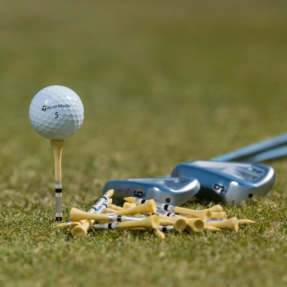 A TaylorMade golf ball positioned on top of a black and gray striped nova bamboo tee with golf clubs beside it. 