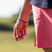 A hand holding a pink pride performance golf tee, with a bracelet on the wrist, against a blurred outdoor background.
