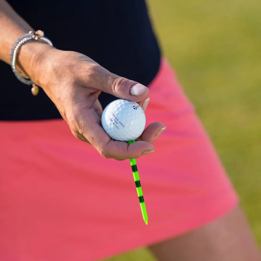 A hand holding a golf ball and tee with a green and black striped design, set against a blurred grassy background.