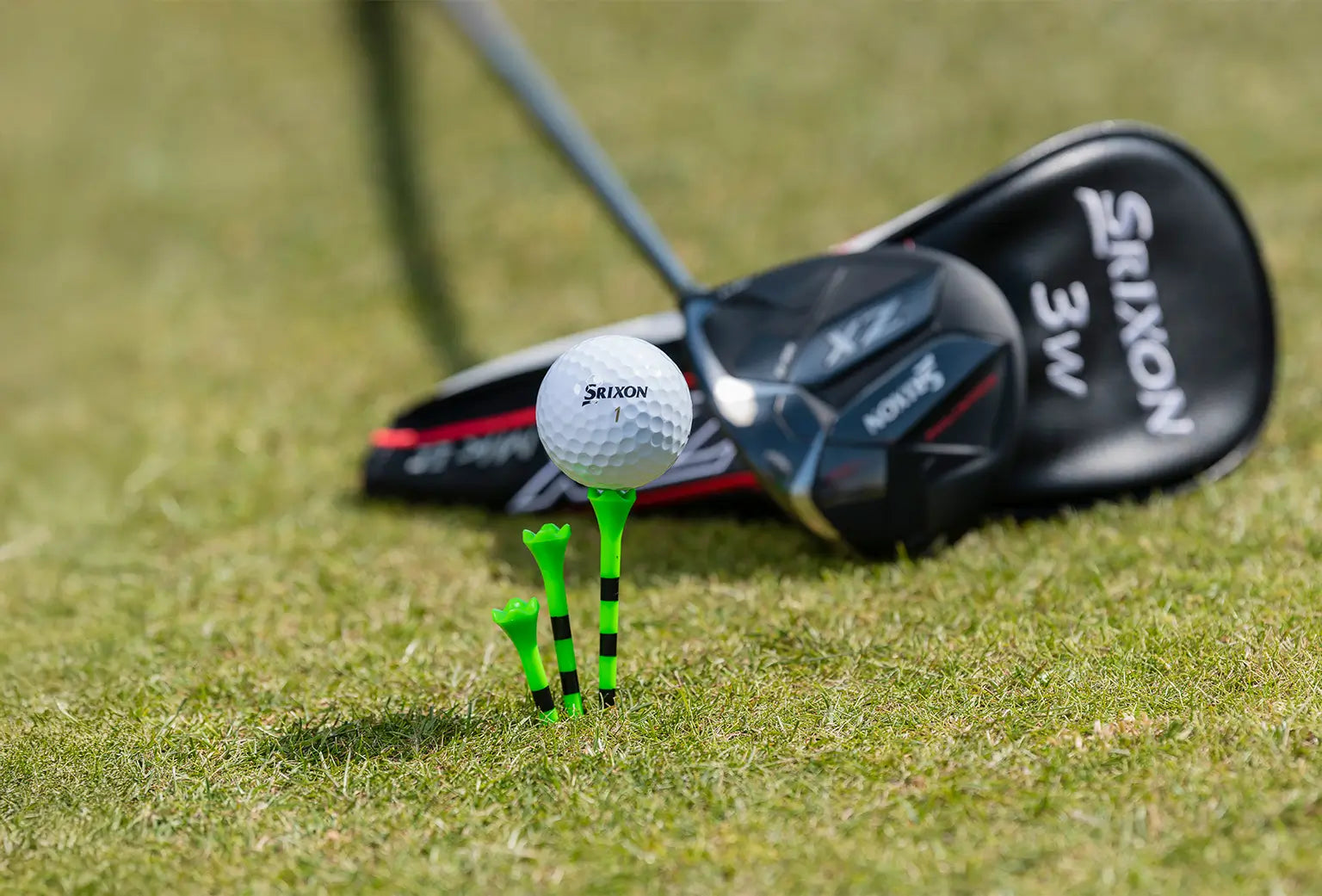 A golf ball positioned on top of a green and black striped golf tee next to two other tees and golf clubs behind it. 