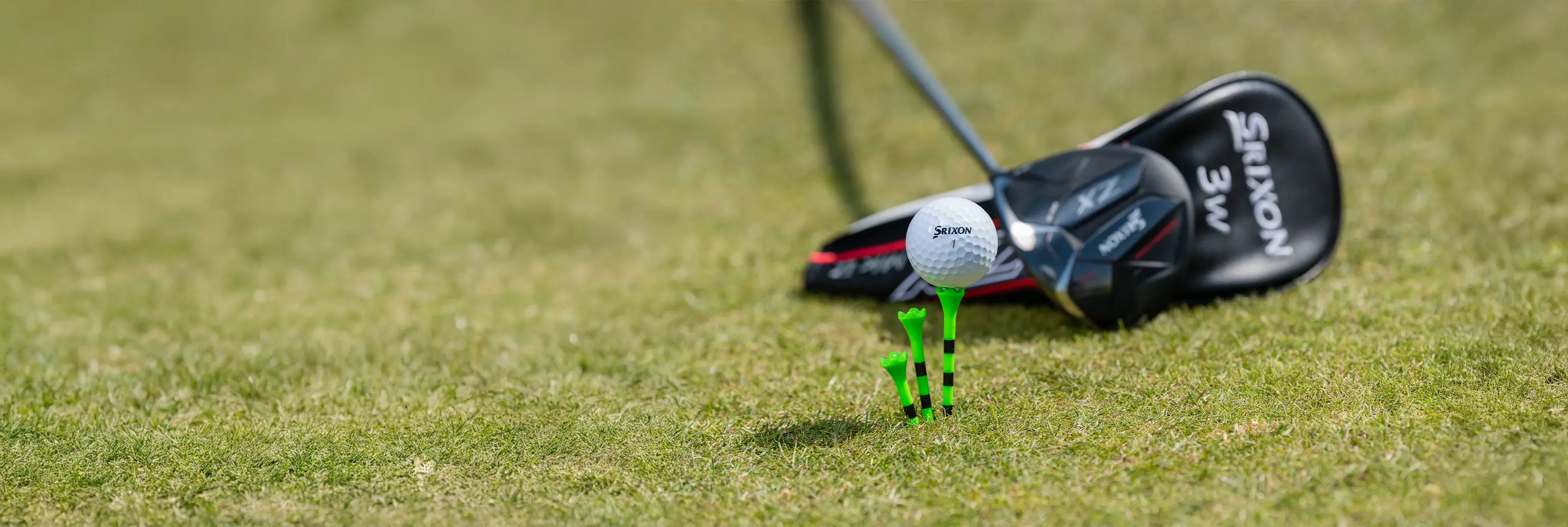 A golf ball positioned on top of a green and black striped golf tee next to two other tees and golf clubs behind it. 