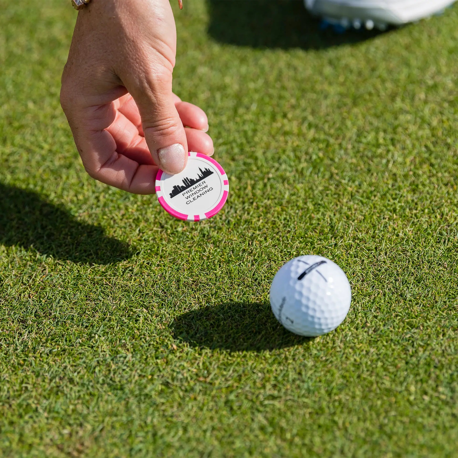 A person holding a pink custom ball marker setting it next to a golf ball on the ground. 