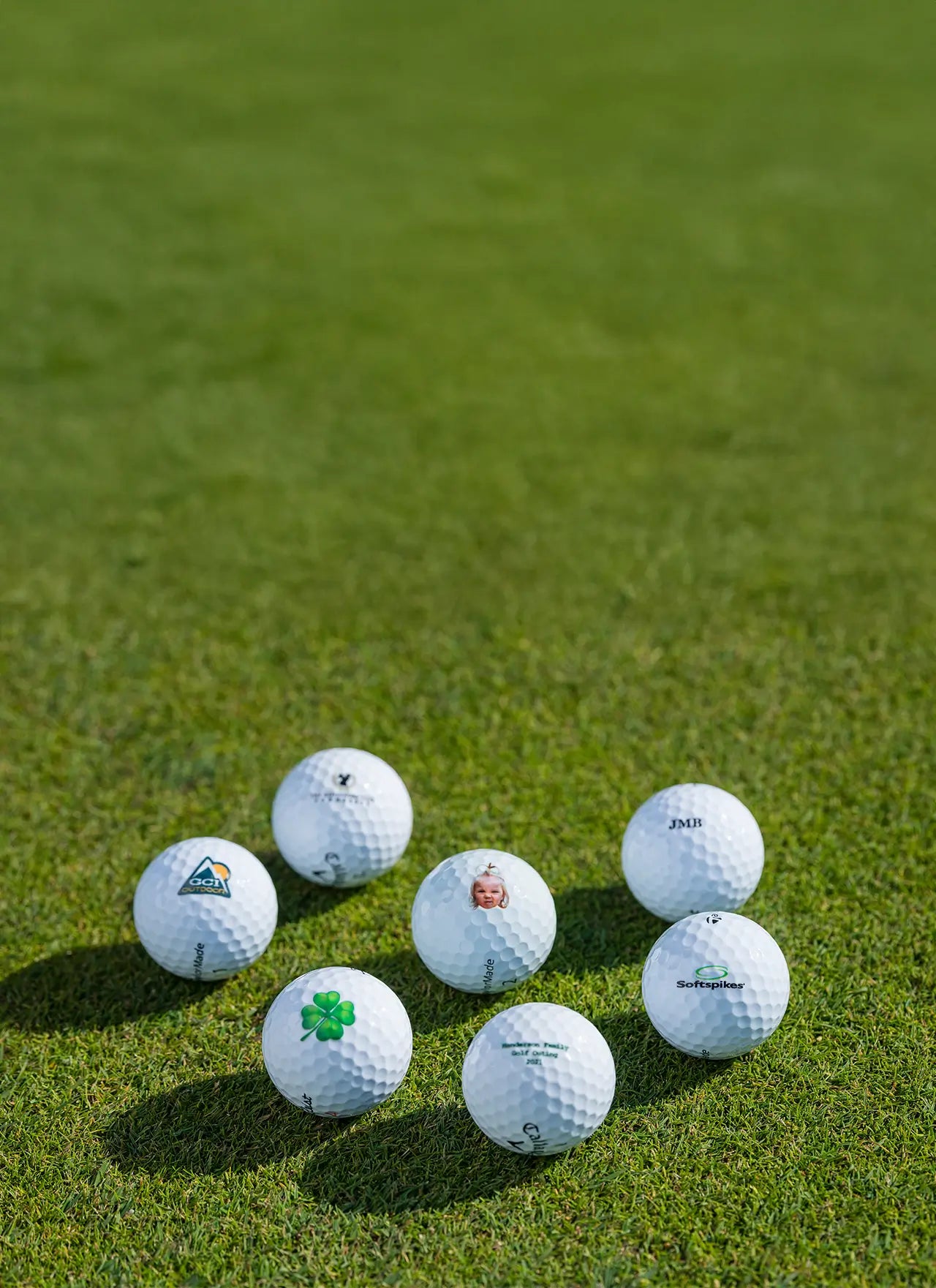 Seven custom golf balls with different logo imprints positioned on green turf. 