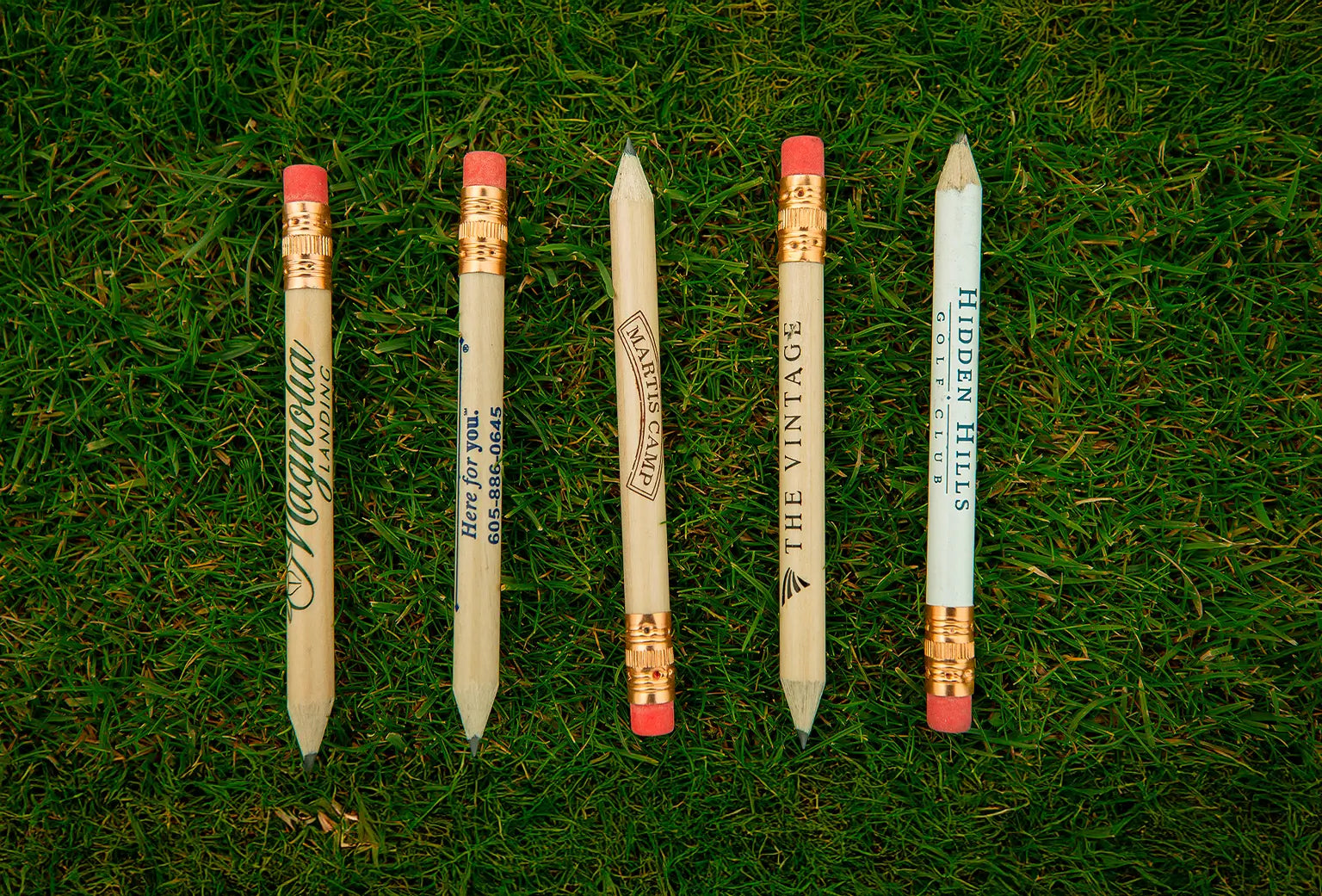 Five custom imprinted golf pencils with different text sitting on green turf. 