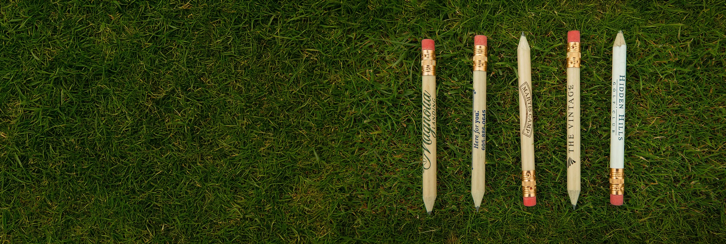 Five custom imprinted golf pencils with different text sitting on green turf. 