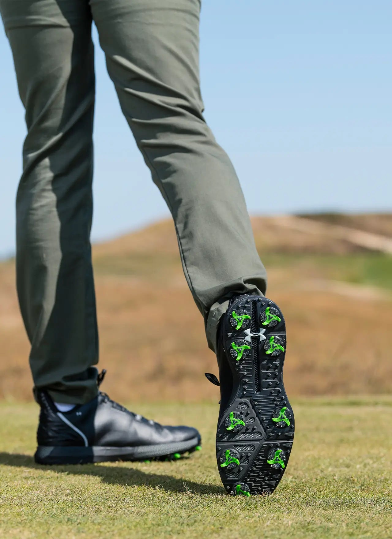 A man showing the bottom of his black under armour shoe with his tour flex pro golf spikes on. 