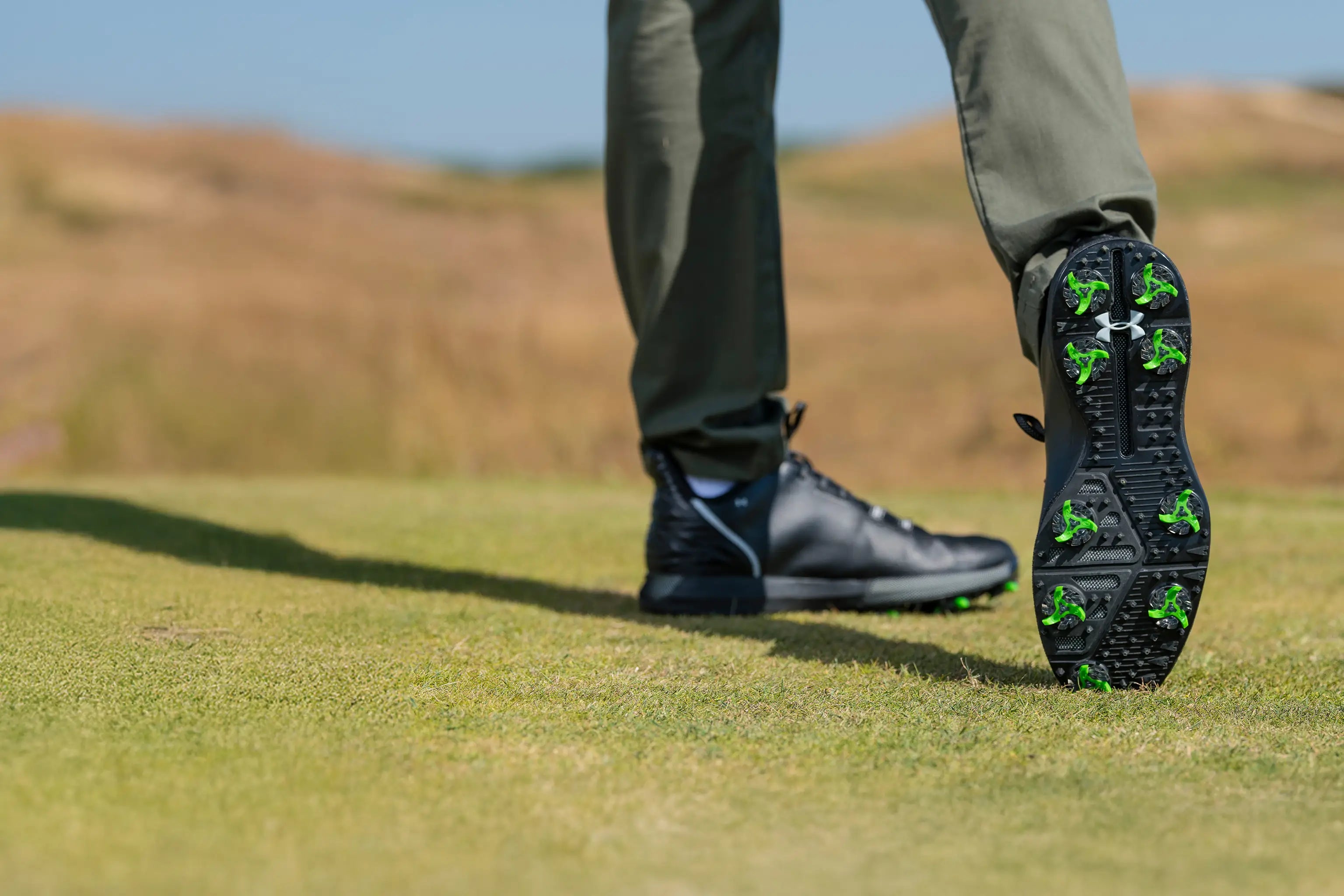 A man showing the bottom of his black under armour shoe with his tour flex pro golf spikes on. 
