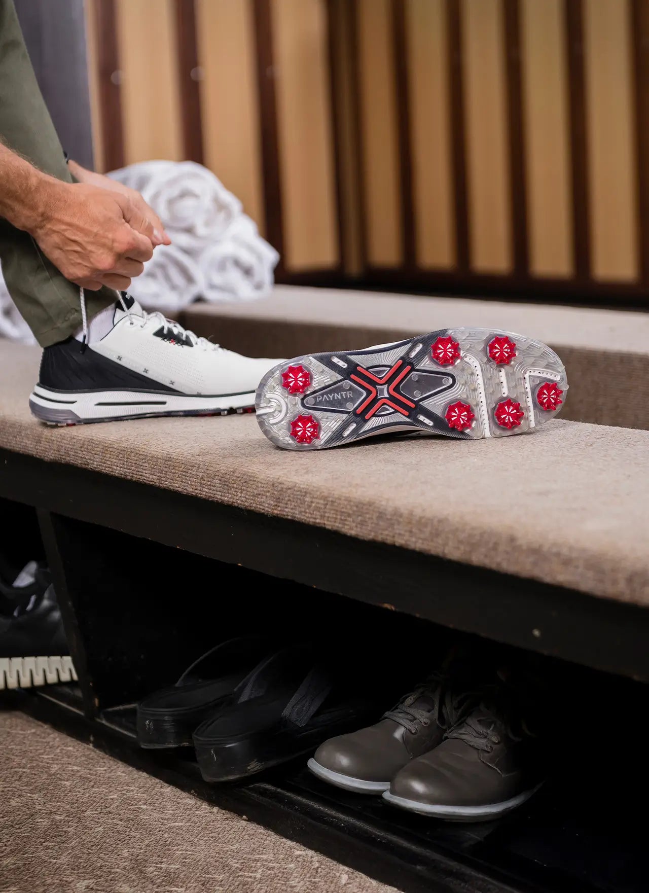A man tying his shoe while his other shoe is positioned upright showing the red pulsar golf spikes. 