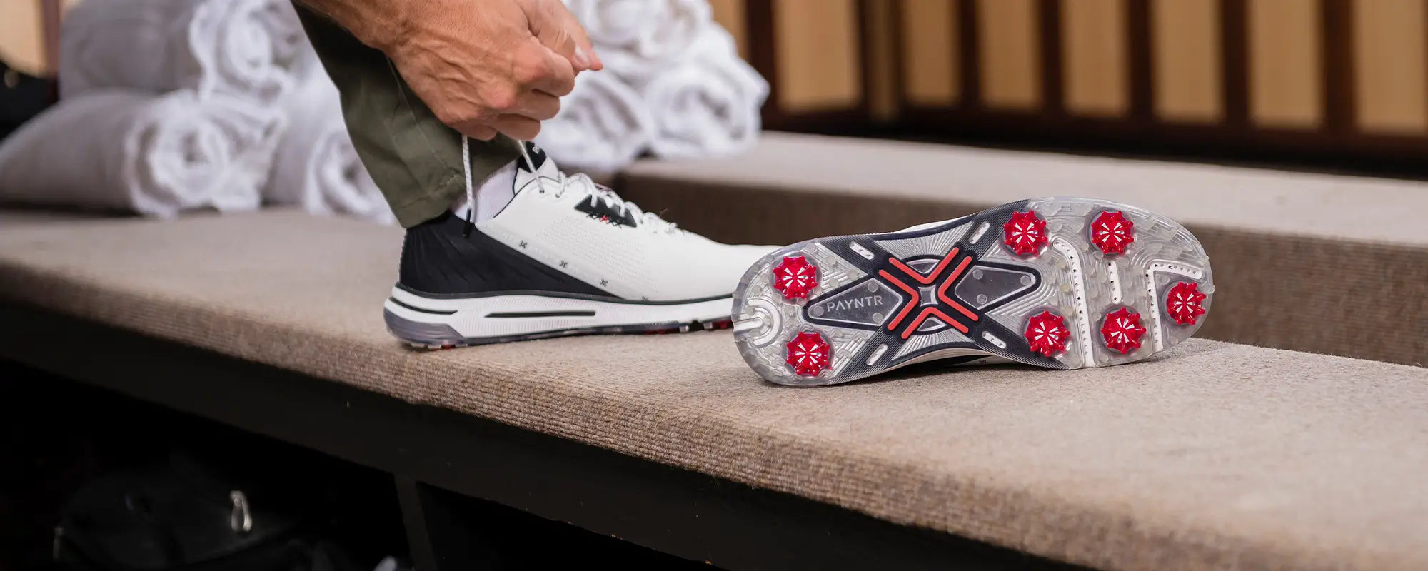 A man tying his shoe while his other shoe is positioned upright showing the red pulsar golf spikes. 