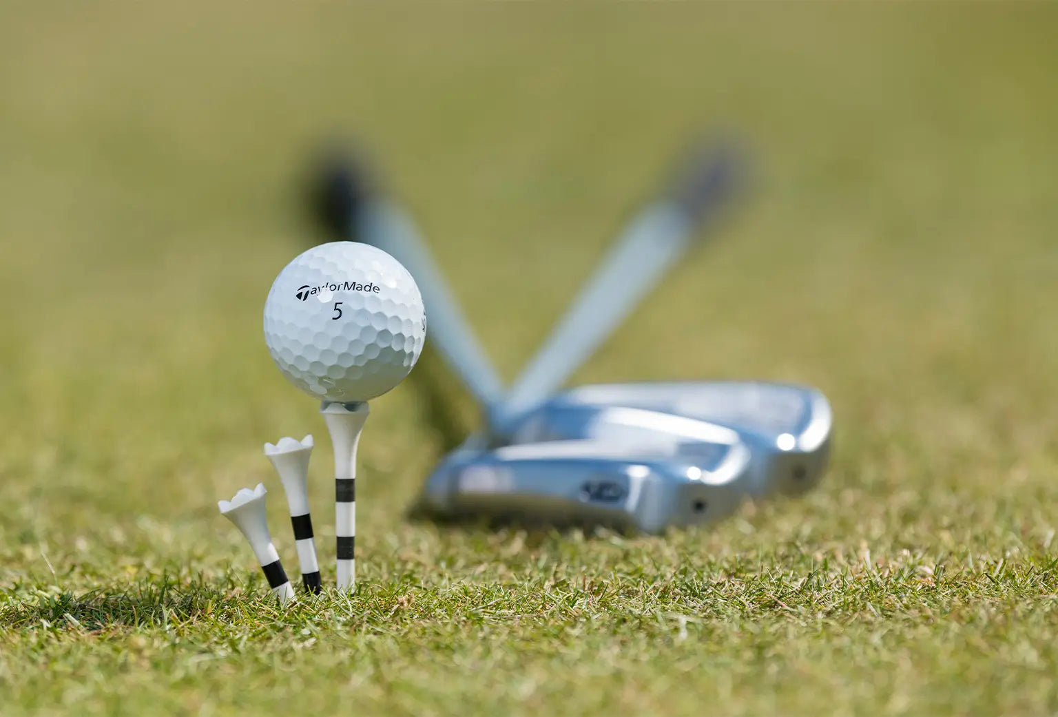 A taylormade golf ball sitting on top of a custom striped pride sports tee with clubs in the background on green turf. 