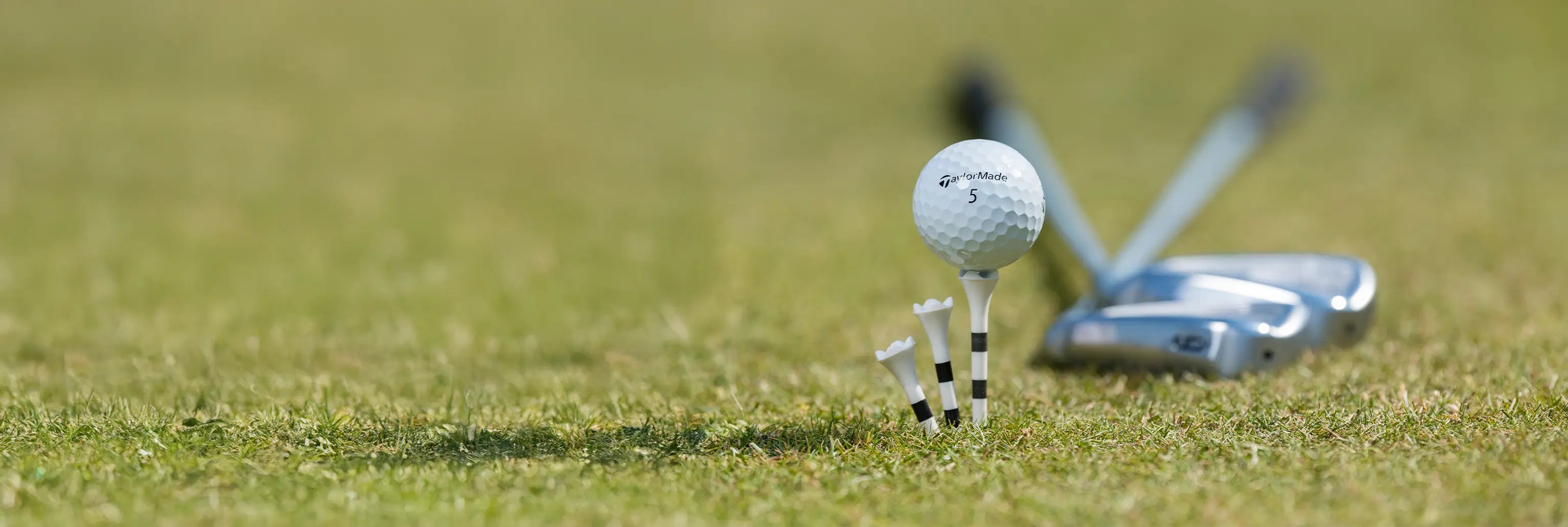 A taylormade golf ball sitting on top of a custom striped pride sports tee with clubs in the background on green turf. 