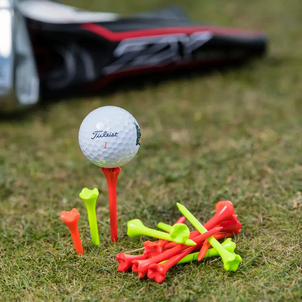 A titleist golf ball on an orange tee with colorful golf tees scattered around on grass, with a golf club partially visible in the background.