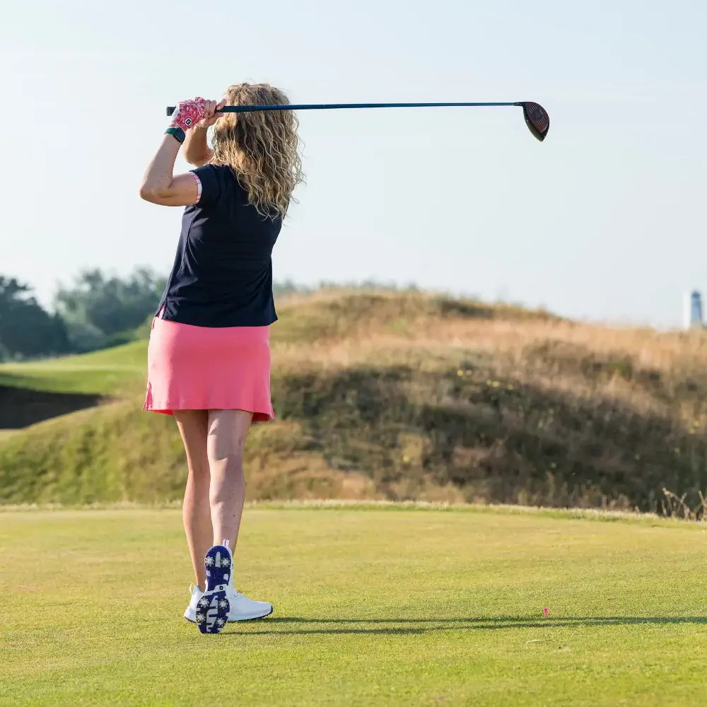 A woman with curly hair is preparing to hit a golf ball on a sunny day, wearing a white shoe featuring Pulsar golf spikes.