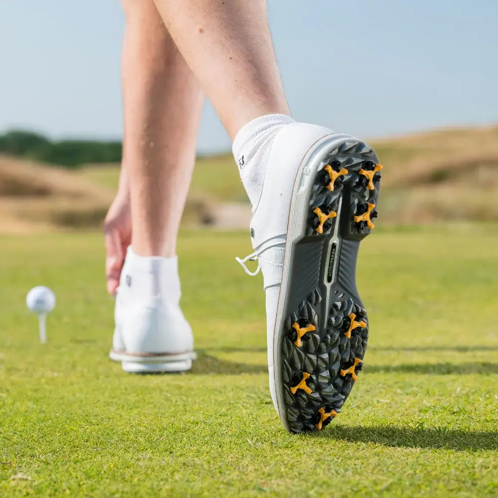 A close-up of a golfer's foot in a white shoe with scorpion stinger golf spikes, standing on green grass near a golf ball on a tee.