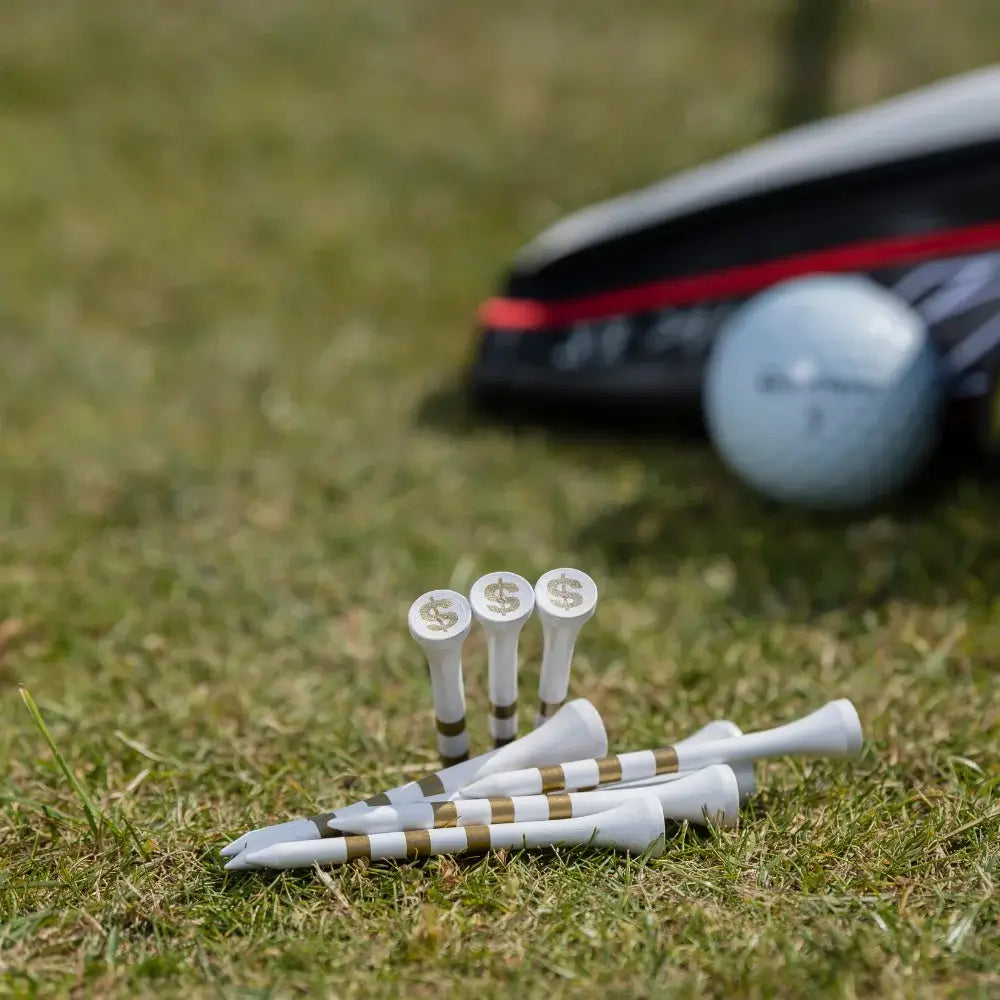 A close-up of golf tees with dollar sign markings, placed on grass, with a golf ball and golf club in the background.