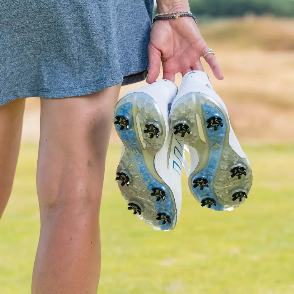 A person holding a pair of white athletic shoes with stealth golf spikes, wearing a gray dress.