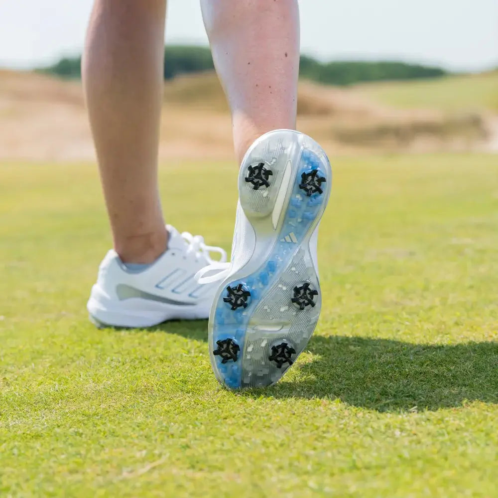 A close-up of a white golf shoe with stealth golf spikes, resting on green grass.