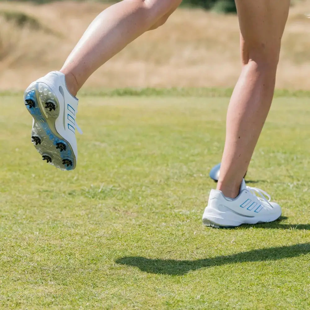 A close-up of a foot wearing a white athletic shoe with stealth golf spikes, stepping on grass.