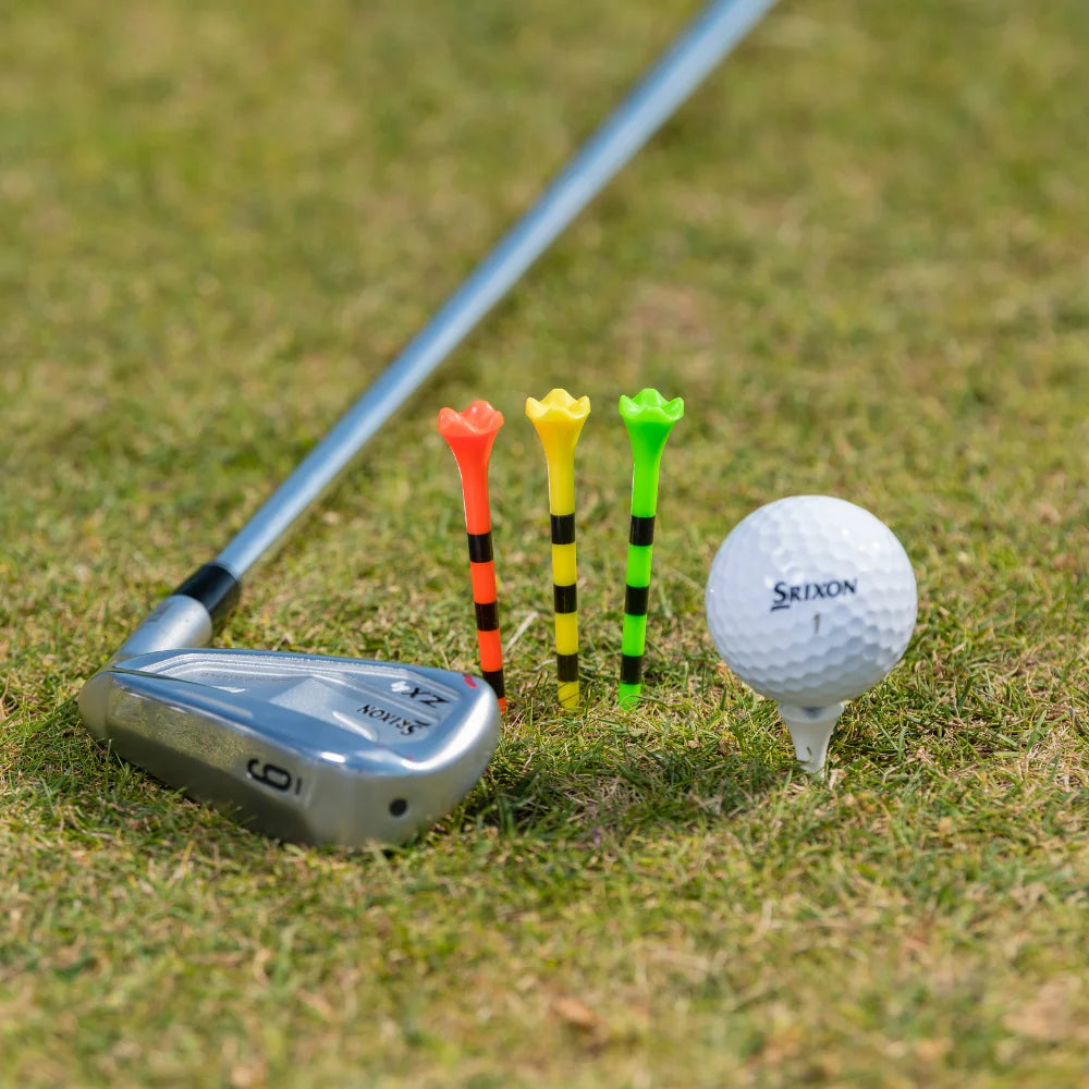 A golf club, three colorful tees (red, yellow, and green with black stripes), and a white srixon golf ball on a tee on grass.
