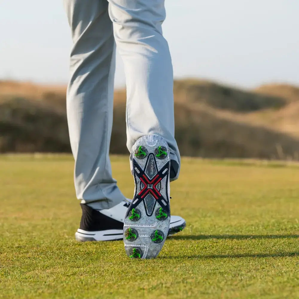 A close-up of a tour flex pro golf spikes on a shoe, with the foot in motion and wearing light-colored pants on a grass.