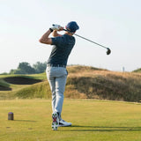 A golfer in a patterned shirt and grey pants swings a driver on a golf course under clear skies, wearing green tour flex pro golf spikes.