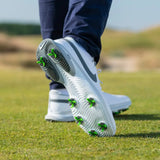 A close-up of a foot wearing a white shoe with green tour flex pro spikes, standing on grassy terrain.