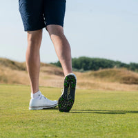 A close-up of a person's legs and golf shoes with green tour flex pro spikes walking on a grassy surface.