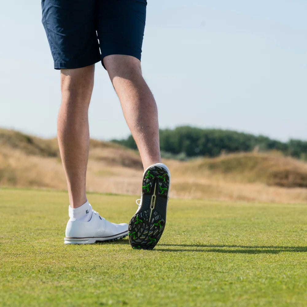 A close-up of a person's legs and golf shoes with green tour flex pro spikes walking on a grassy surface.