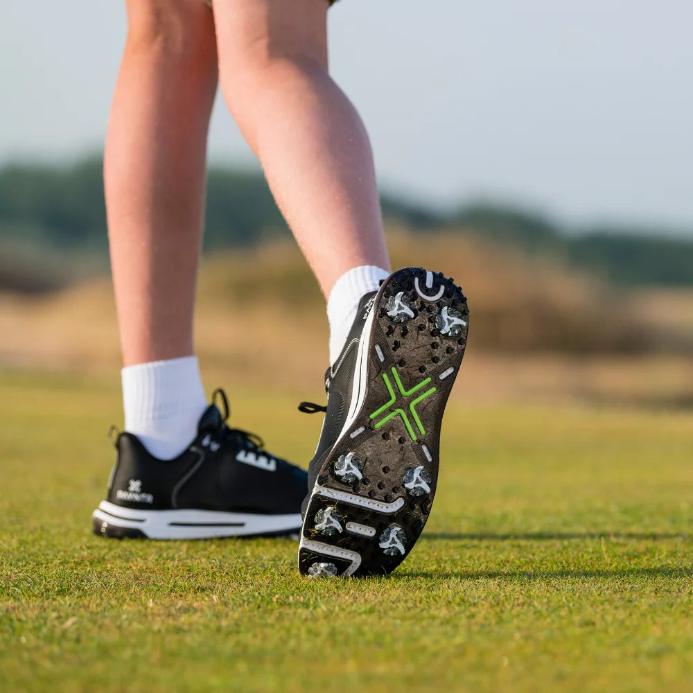 A close-up of a foot wearing a black shoe with green tour flex pro spikes, standing on grassy terrain.