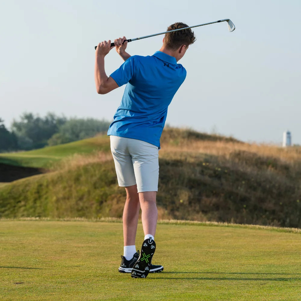 A young golfer in a blue polo shirt,  white shorts, and black shoe with white spikes is swinging a golf club on a grassy course.