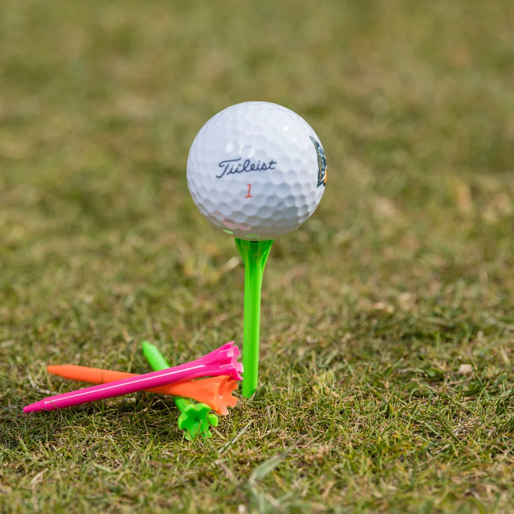 A titleist golf ball on a green tee, with additional colored tees (orange, pink, and green) scattered nearby on grass.