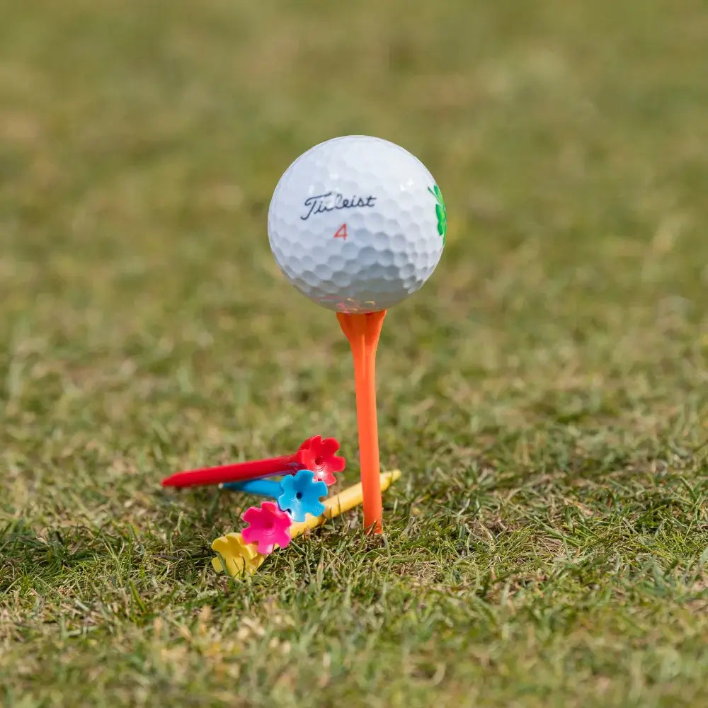 A white titleist golf ball on an orange tee, with colorful plastic golf tees in red, blue, pink, and yellow resting on the grass.